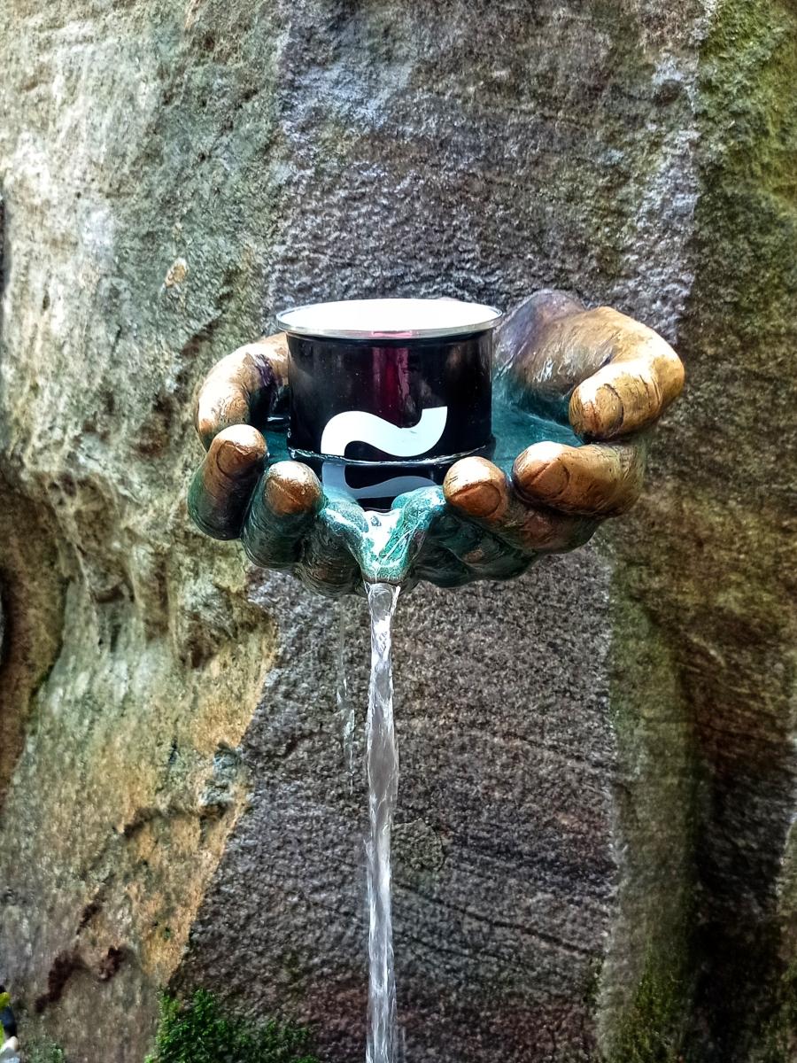 Skalní pramen s výtokem ve tvaru rukou s nabranou vodou, v rukou je plecháček s jabockým logem a pod ním vytéká voda