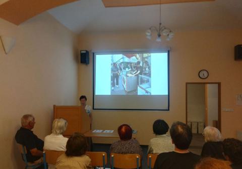 Návštěvníci sledují přednášku Pohled zedadu na návštěvníky akce, kteří sledují přednášející a obrázek pouličního prodavače na promítacím plátně.