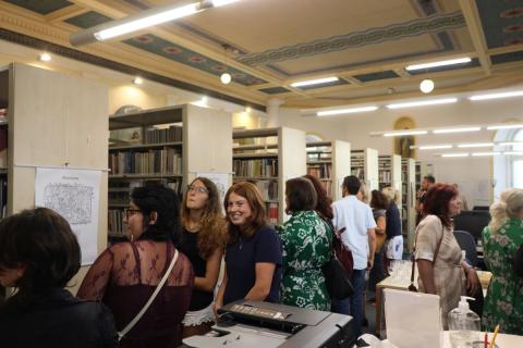 Návštěvníci vernisáže Prostor knihovny plný lidí, kteří spolu hovoří, někteří si prohlížejí obrázky zavěšené an boku regálů.