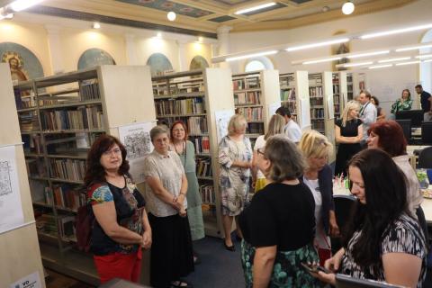 Návštěvníci vernisáže při rozhovorech a prohlídce obrázků Prostor knihovny plný lidí, kteří spolu hovoří, někteří si prohlížejí obrázky zavěšené an boku regálů.