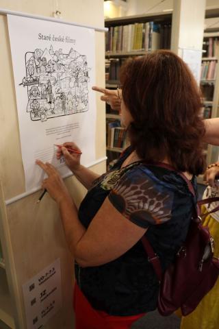 Interakce návštěvnice s vystavenými obrázky Dívka stují u boku regálu, na kterém je zavěšen obrázek a píše na něj