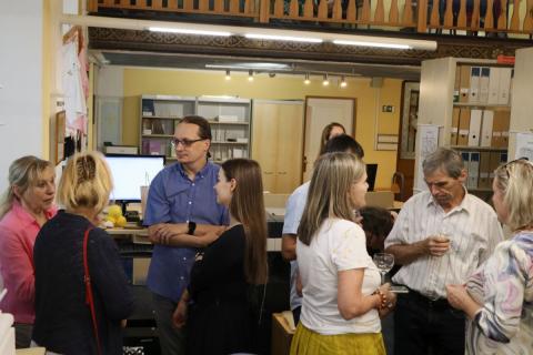 Návštěvníci vernisáže při rozhovorech Skupinky lidí stojící velle regálů a výpůjčního pultu v knihvně, kteří spolu hovoří, někteří drží sklenici s nápojem