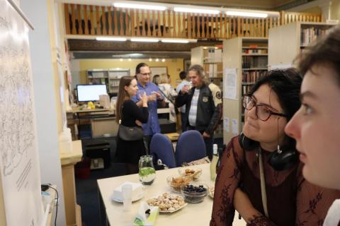 Návštěvnicehledají pojmy na vystaveném obrázku Obličeje dvou dívek zkoumající vystavený obrázek, vzadu je vidět stůl s občerstvením a další návštěvníci akce