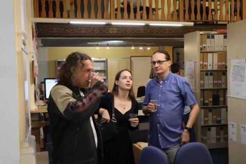 Návštěvníci vernisáže v rozhovoru Dva muži a dívka se spolu povídají, jeden z mužů pije ze sklenice, za skupinou jsou vidět vystavené obrázky na boku regálu a další návštěvníci