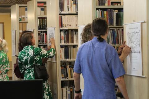 Interakce návštěvníků s vystavenými obrázky U boků dvou regálů na kterých visí vystavené obrázky stojí lidé a vybarvují obrázky