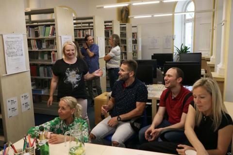 Návštěvníci vernisáže v družné zábavě Skupina lidí sedí v knihovně u stolu s jídlem a povídají si a smějí se. Za nimi jsou vidět další lidé, kteří si povídají u regálů s vystavenými obrázky