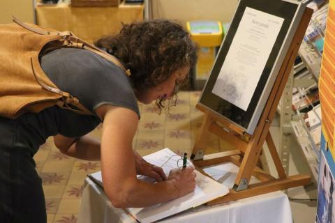 Zápis do návštěvní knihy Žena se sklání u stolečku s plakátem k výstavě a návštěvní knihou, do které něco píše.