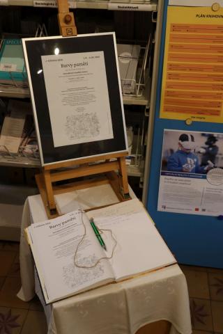 Návštěvní kniha Na stolečku stojí malířský stojan se zarámovaným plakátem, pod ním leží návštěvní kniha a pero